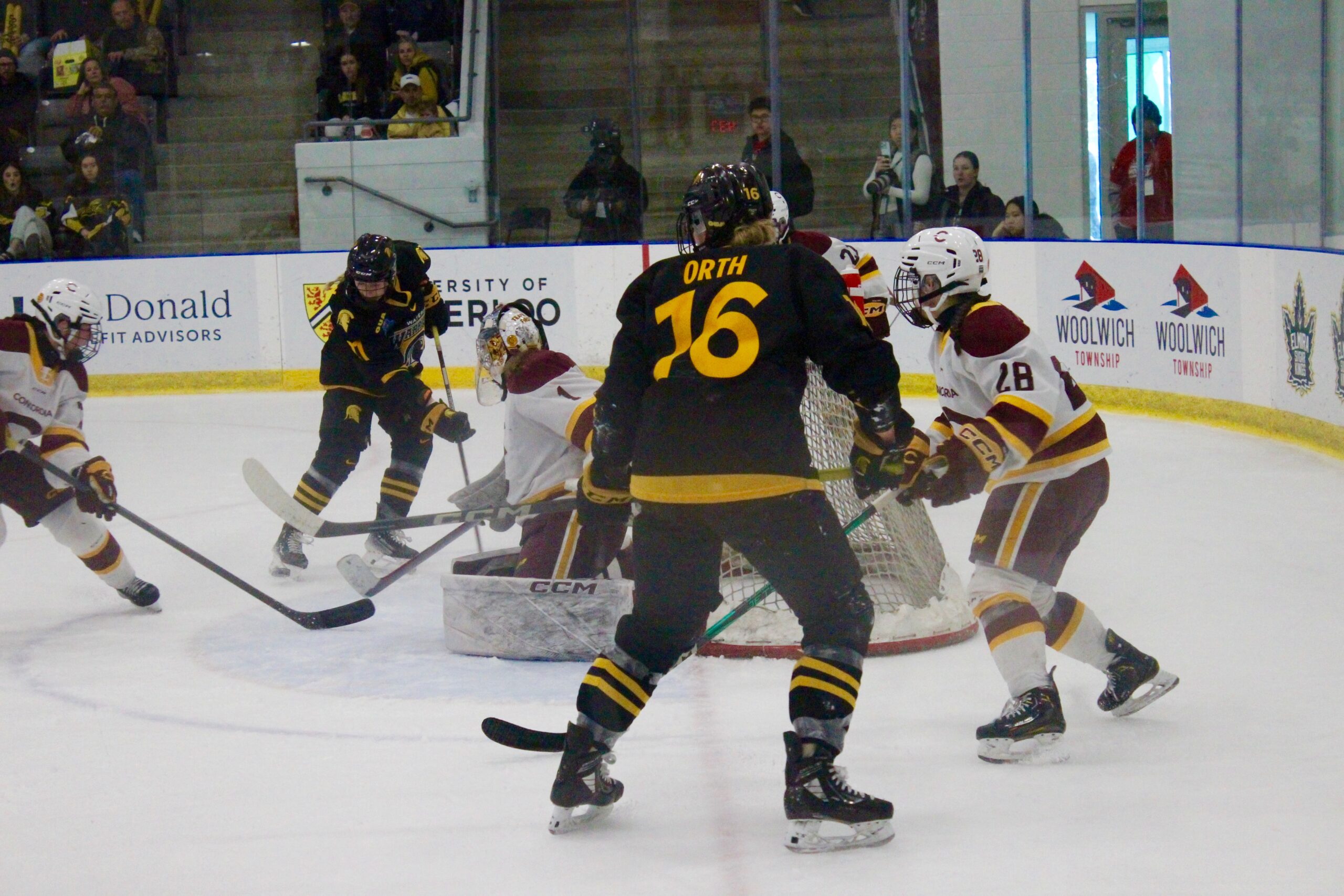 Waterloo's Kassidy McCarthy in black and yellow uniform scores the third goal against Concordia players in white and maroon near the goal. Spectators watch from the stands.