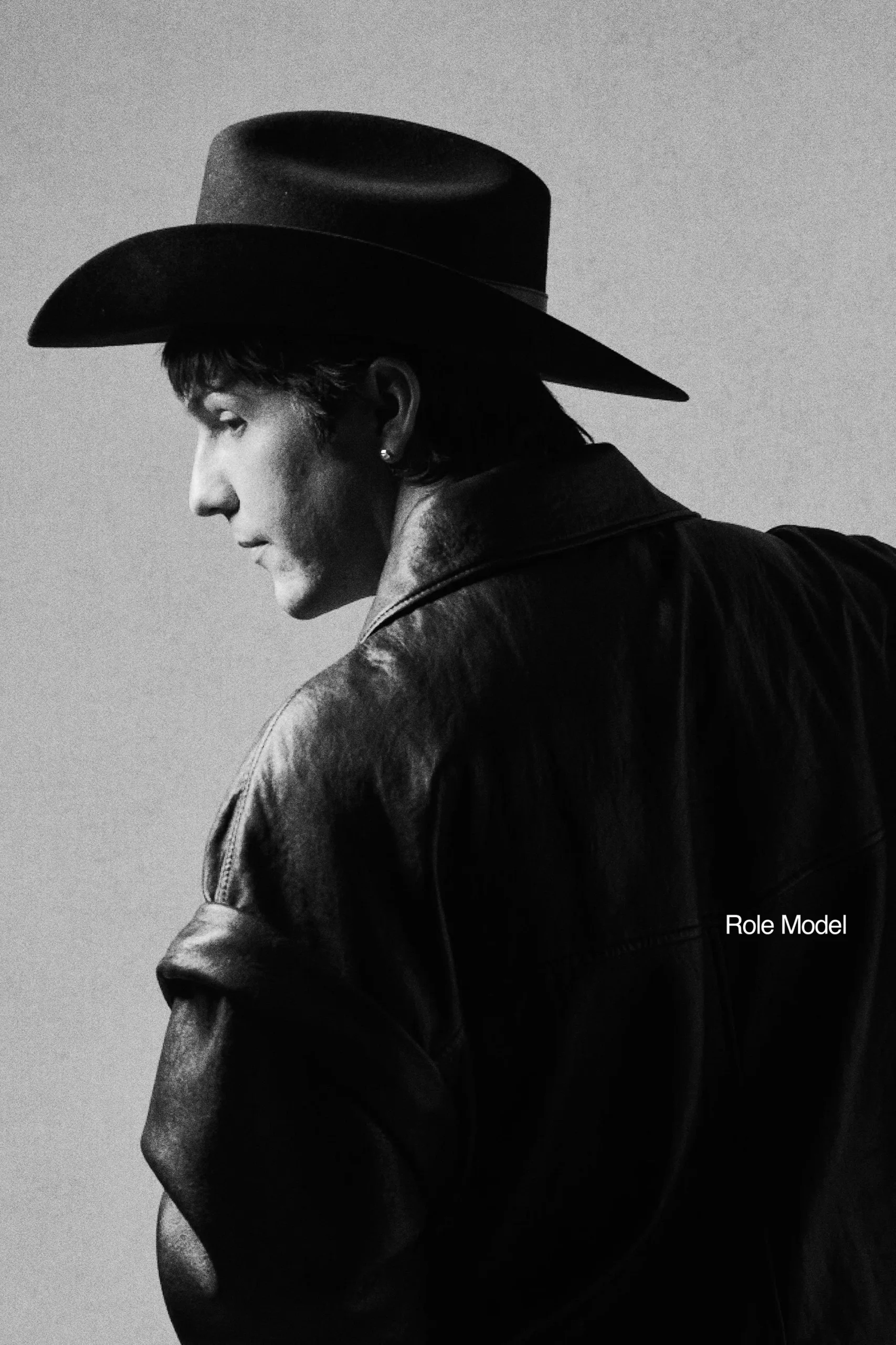 Black and white image of a person in a wide-brimmed hat and leather jacket, facing slightly away. The background is plain, and the text "Role Model" is visible on the jacket. The lighting creates soft shadows.