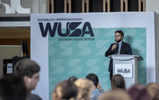 A man in a suit stands at a podium with a microphone, addressing an audience. The backdrop displays "Waterloo Undergraduate Student Association" and "WUSA." People are seated in the foreground, listening attentively as he discusses the winter annual meeting agenda.