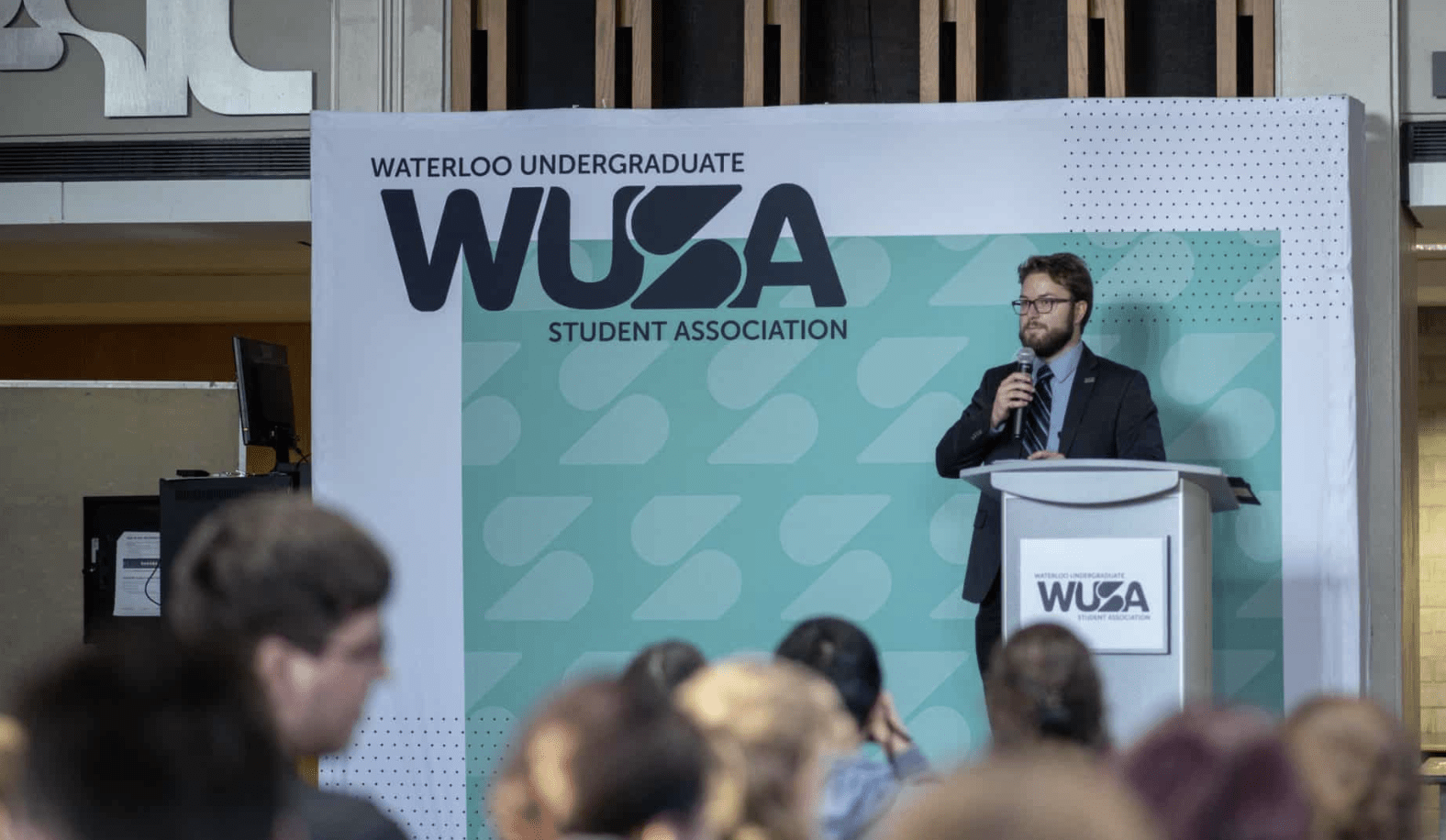 A man in a suit stands at a podium with a microphone, addressing an audience. The backdrop displays "Waterloo Undergraduate Student Association" and "WUSA." People are seated in the foreground, listening attentively as he discusses the winter annual meeting agenda.