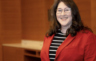 Whitney Barrett, wearing glasses and a red blazer over a black and white striped top, smiles in a warmly lit room with wooden walls, embodying the approachable presence of an UWaterloo Ombudsperson.