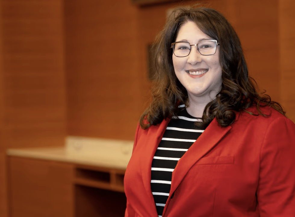 Whitney Barrett, wearing glasses and a red blazer over a black and white striped top, smiles in a warmly lit room with wooden walls, embodying the approachable presence of an UWaterloo Ombudsperson.