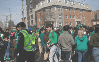 A large outdoor gathering of people, many wearing green clothing, fills a street in the City of Waterloo in front of brick buildings. Two police officers in yellow vests stand by. The scene suggests a festive event, likely St. Patrick's Day celebrations.