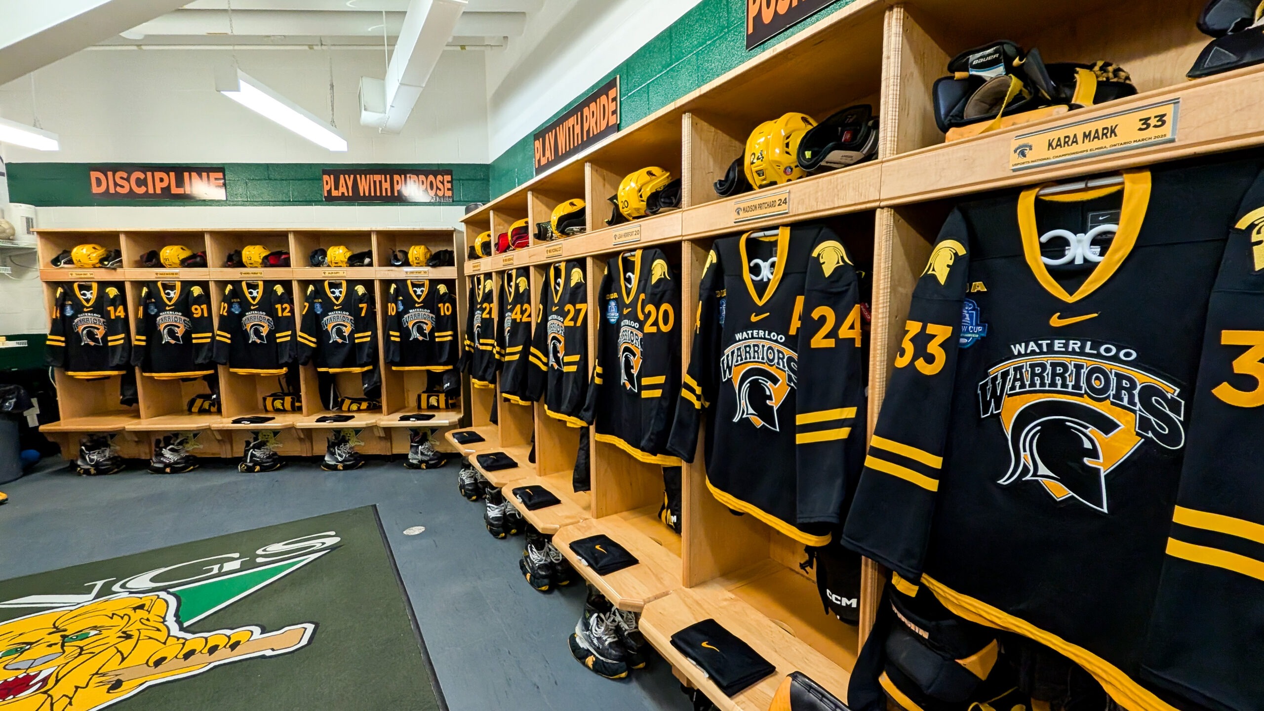A locker room with a row of black and yellow hockey jerseys, proudly displaying "Waterloo Warriors," hangs in open wooden cubbies. Each jersey hints at the fierce battles of the 2024-2025 women's hockey season. Motivational phrases inspire from above.