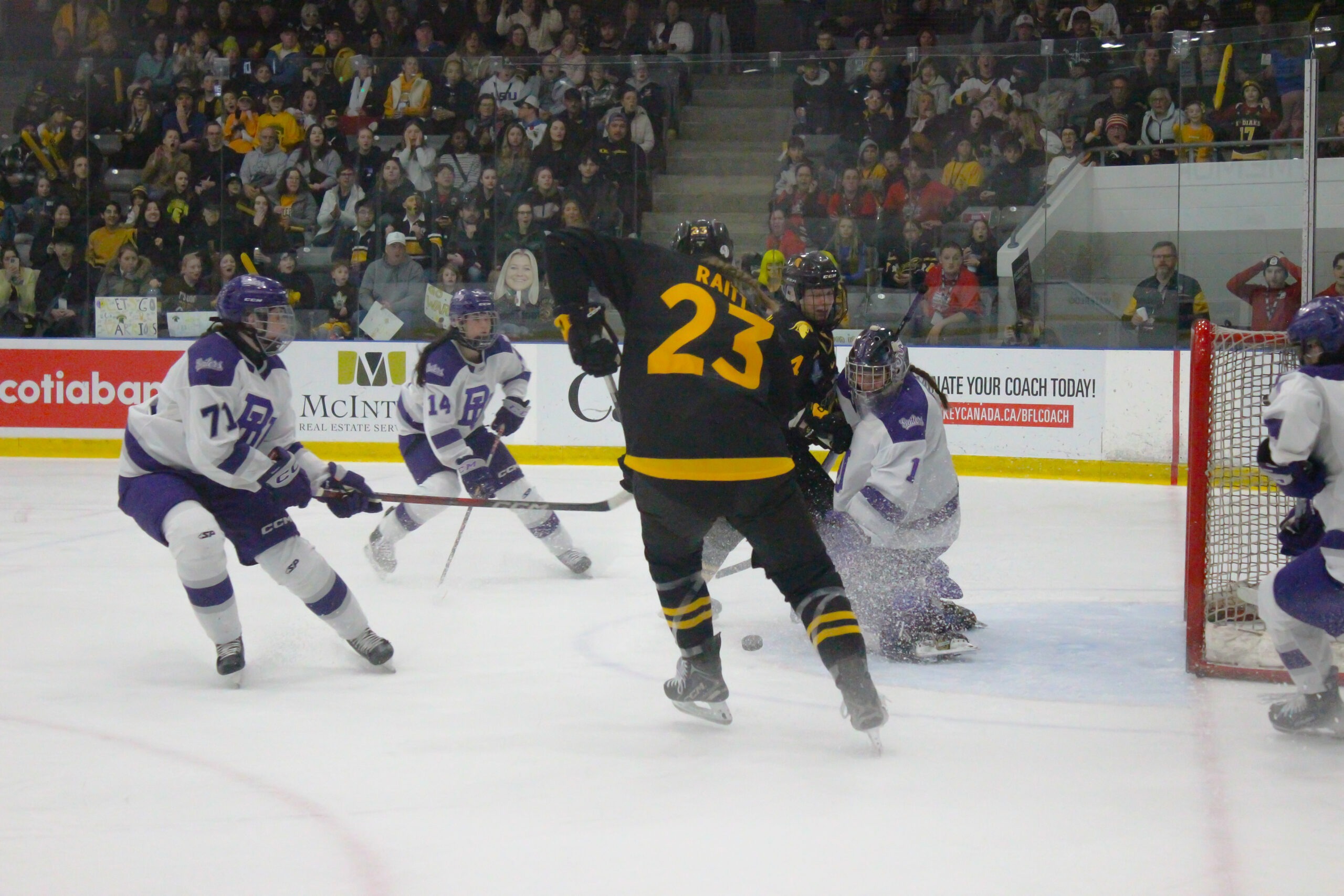 A hockey player wearing a black jersey with number 23 from the Warriors women's hockey team attempts to score a goal against the opposing team in purple and white uniforms. The goalie braces to block the shot as spectators watch eagerly, recalling past U Sports silver victories.