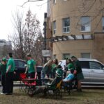 A group of people in green clothing stand and sit near a building and parked cars, socializing outdoors. Some are wearing festive accessories. Foldable chairs and a table are set up on the grass. Urban buildings are visible in the background.