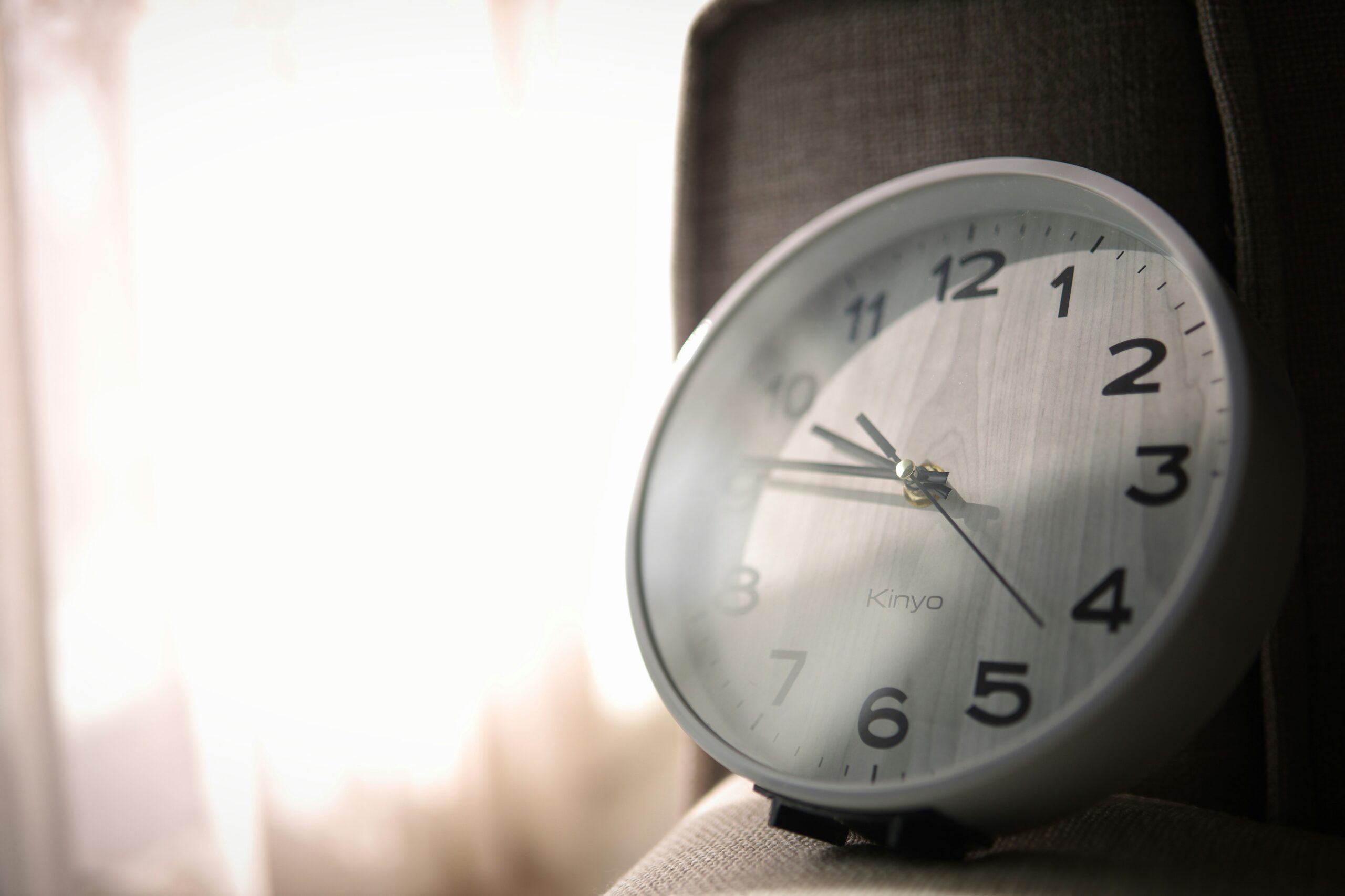 A white analog clock with black numbers and hands rests on a cushioned surface at an angle, adjusting to the time change. Sunlight filters through sheer curtains in the background, creating a soft, warm glow.