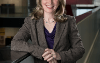 A person with long hair, presumably Dean of Faculty Mary Wells, smiles while wearing a business suit and purple blouse, leaning on a railing in the modern engineering building's interior. Stairs and glass walls elegantly frame the background.