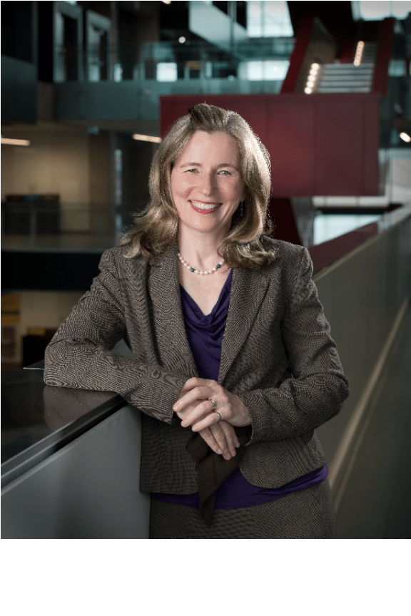 A person with long hair, presumably Dean of Faculty Mary Wells, smiles while wearing a business suit and purple blouse, leaning on a railing in the modern engineering building's interior. Stairs and glass walls elegantly frame the background.