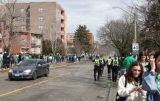 A crowd of people, many wearing green, lines a street near apartment buildings during Waterloo St. Patrick's Day celebrations. Several police officers in yellow vests walk down the road. A car is parked nearby and a 40 km/h speed sign is visible.
