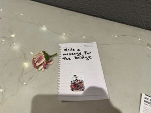 A small notepad rests on a table with "Write a message for the MC-DC bridge" inscribed on the top page. A single pink flower and gentle string lights accompany it, alongside a sticker depicting a group of people, adding warmth to the heartfelt scene.