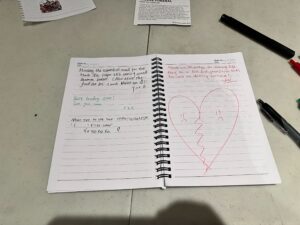 An open spiral notebook rests on the table, filled with handwritten notes and a red heart drawing divided in two. Nearby, two black pens lie beside a folded piece of paper—perhaps remnants of reflection from crossing life's MC-DC bridge.