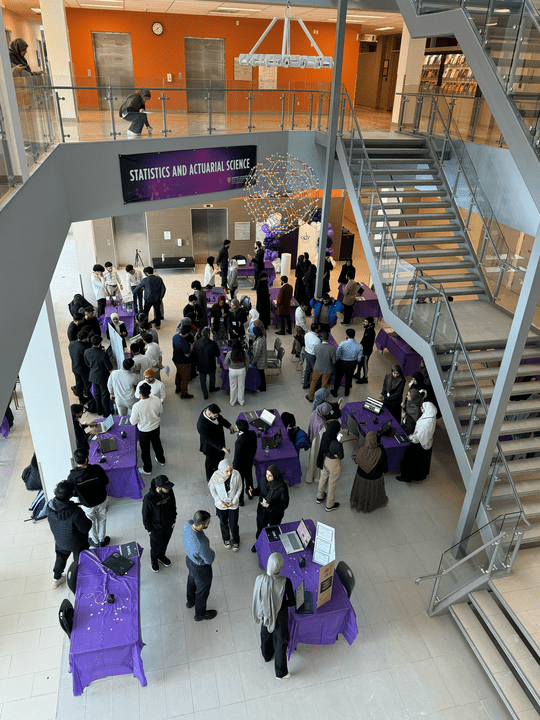 People gather in a multi-level atrium for the UW Statistics and Actuarial Science event. Attendees visit tables draped in purple cloths, while a few signs from the Ibtikar Summit are visible. Stairs and an upper balcony provide a perfect vantage point over the vibrant event space.