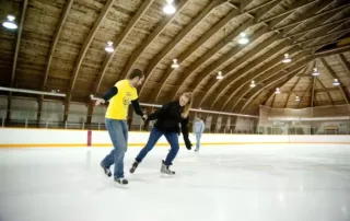 Two people ice skating together inside a wooden-roofed rink, holding hands for balance like a beginner’s guide. A third person skates alone in the background under the arched ceiling with bright lights.