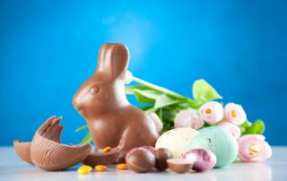 A chocolate bunny sits beside colorful Easter eggs, assorted candies, and pink tulips on a white surface with a bright blue background—perfect for celebrating the Easter long weekend.