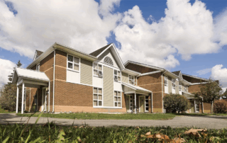 A two-story brick apartment building with white trim, large windows, and a sloped roof sits on a grassy lawn under a partly cloudy sky—ideal guaranteed housing for UWaterloo PhD students. Autumn leaves are scattered on the grass in the foreground.