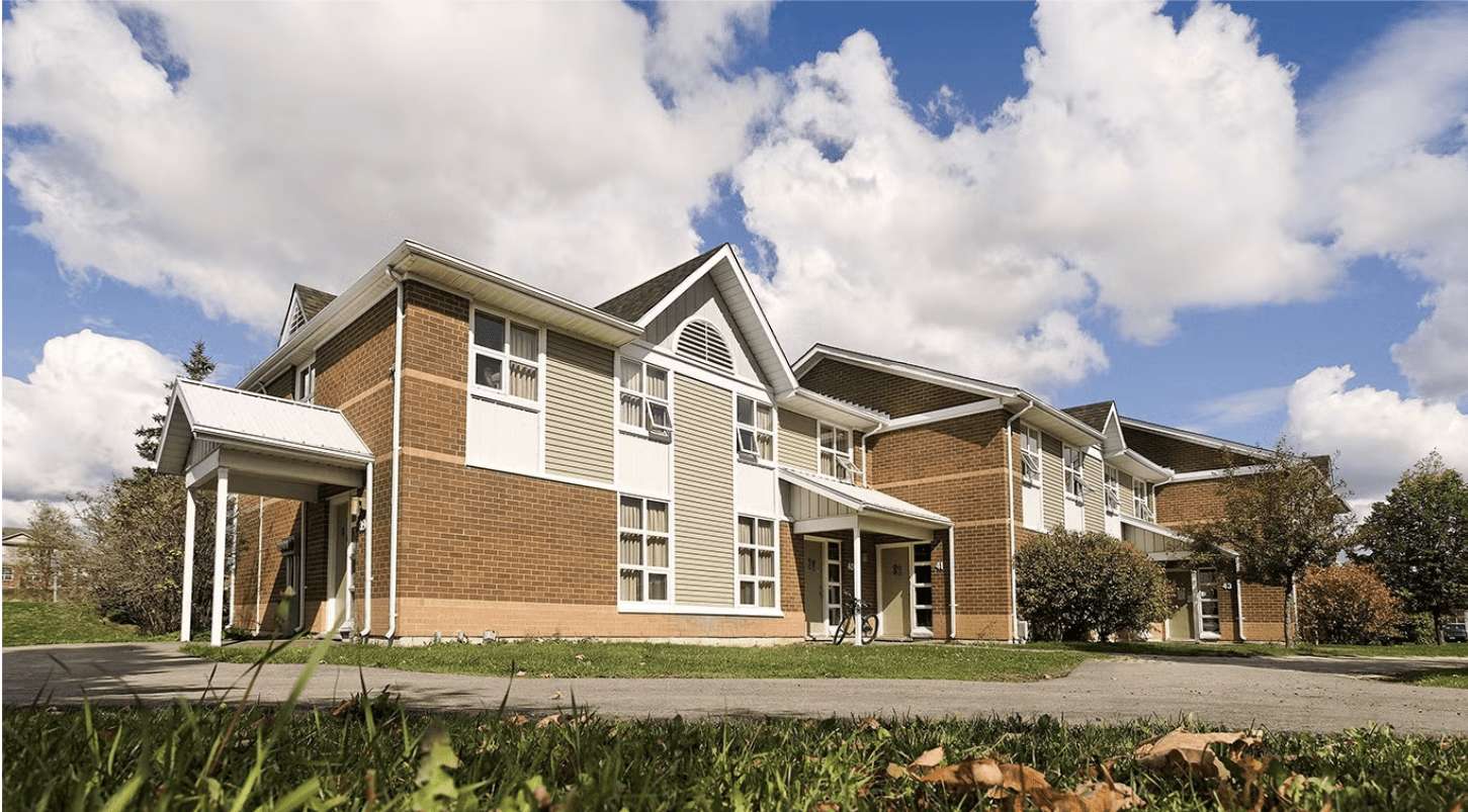 A two-story brick apartment building with white trim, large windows, and a sloped roof sits on a grassy lawn under a partly cloudy sky—ideal guaranteed housing for UWaterloo PhD students. Autumn leaves are scattered on the grass in the foreground.