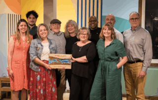 A group of eleven adults, smiling and posing indoors, celebrates 10 years of community. One person in front holds a decorative wooden plaque. The background features colorful, geometric wall art and a window at Seven Shores Café.