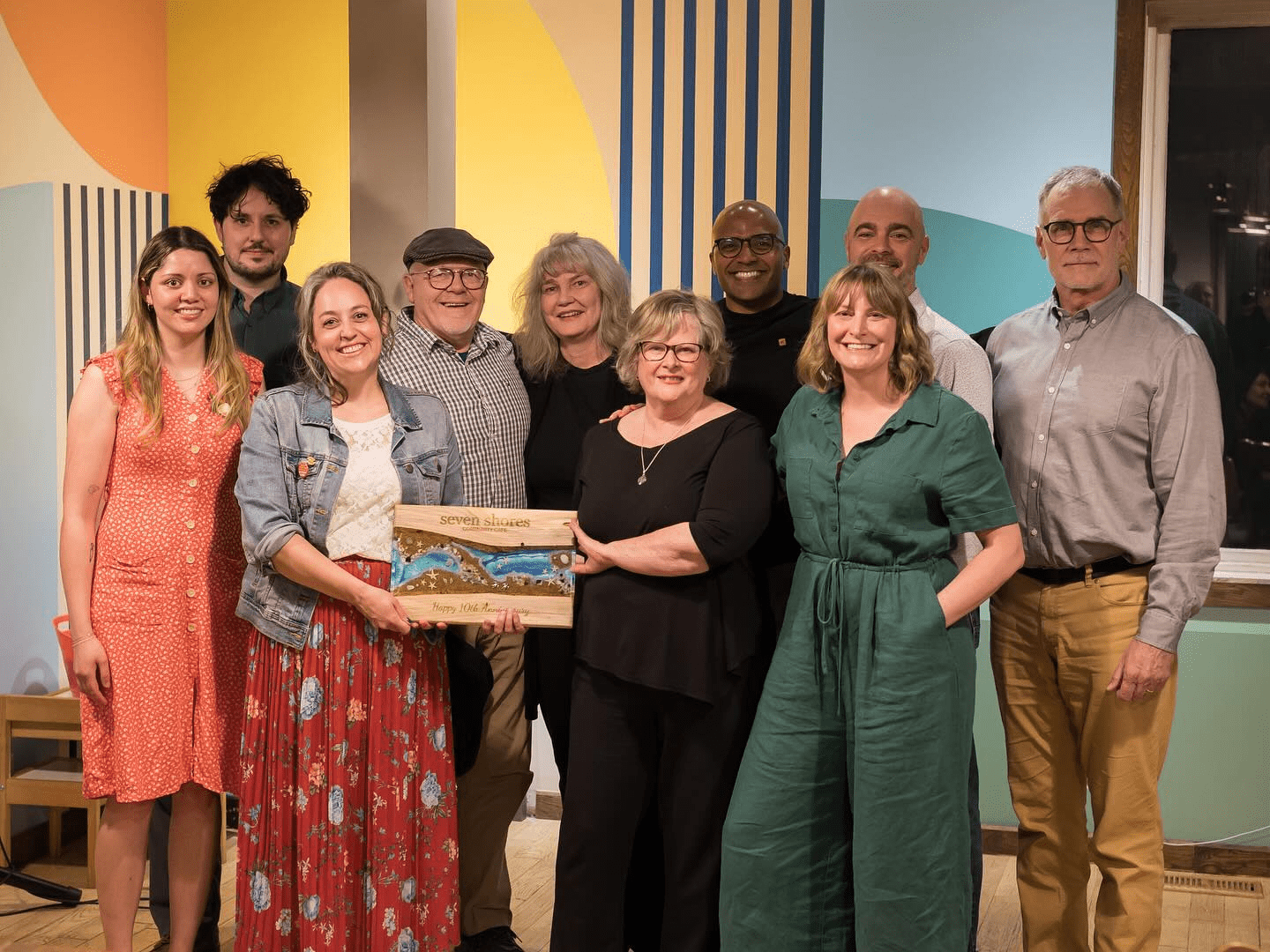 A group of eleven adults, smiling and posing indoors, celebrates 10 years of community. One person in front holds a decorative wooden plaque. The background features colorful, geometric wall art and a window at Seven Shores Café.