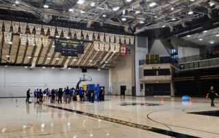 A group of people in blue shirts stands in a line on an indoor basketball court, with RBC Training Ground banners hanging from the ceiling and empty bleachers in the background. Orange cones mark areas on the court.