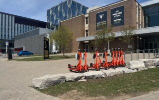 A row of orange electric scooters is parked near a UW building with large windows and brick walls. Nearby, there are trees, a digital sign, and a blue car—signs of campus life returning for the spring term.