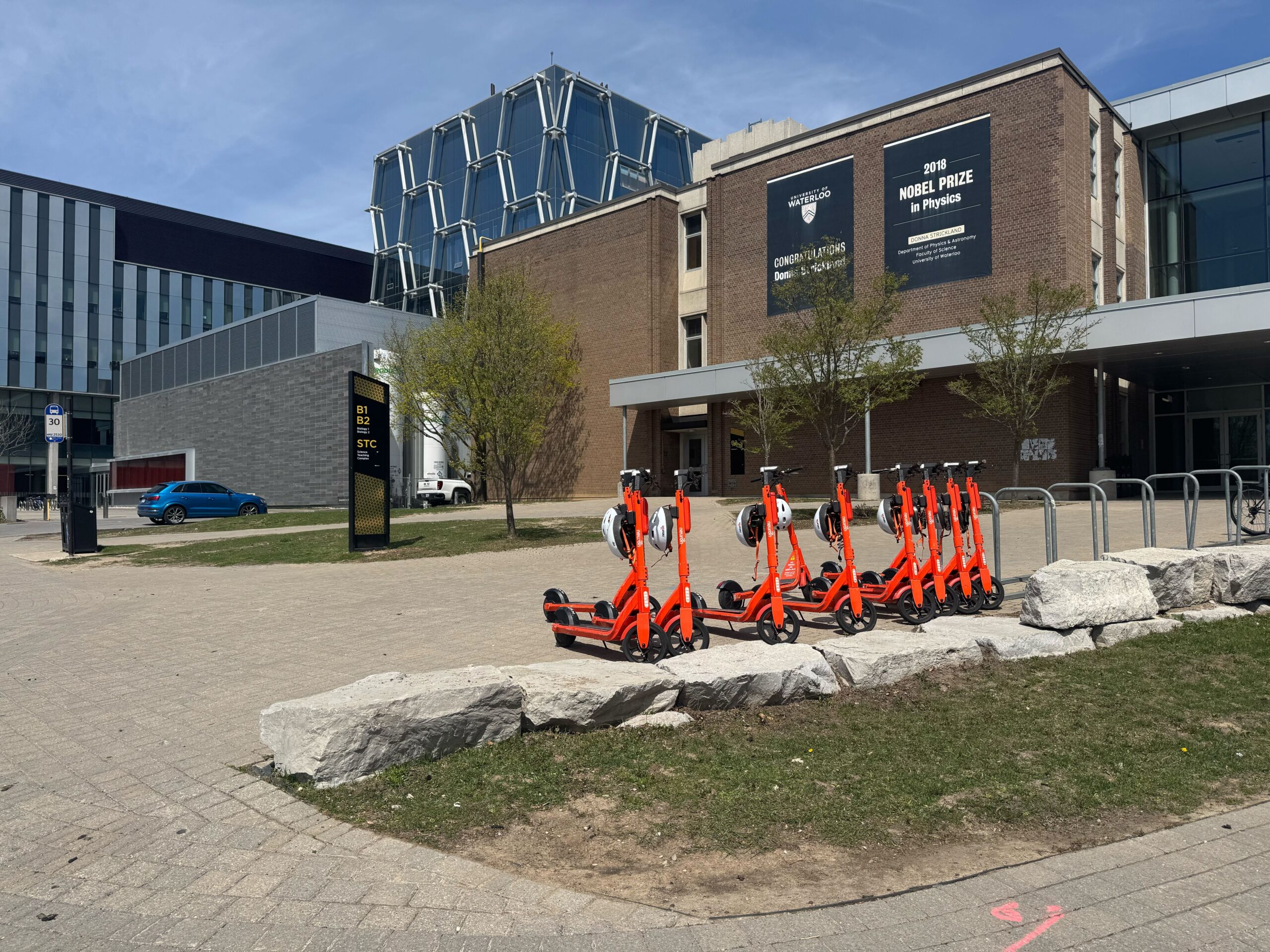 A row of orange electric scooters is parked near a UW building with large windows and brick walls. Nearby, there are trees, a digital sign, and a blue car—signs of campus life returning for the spring term.