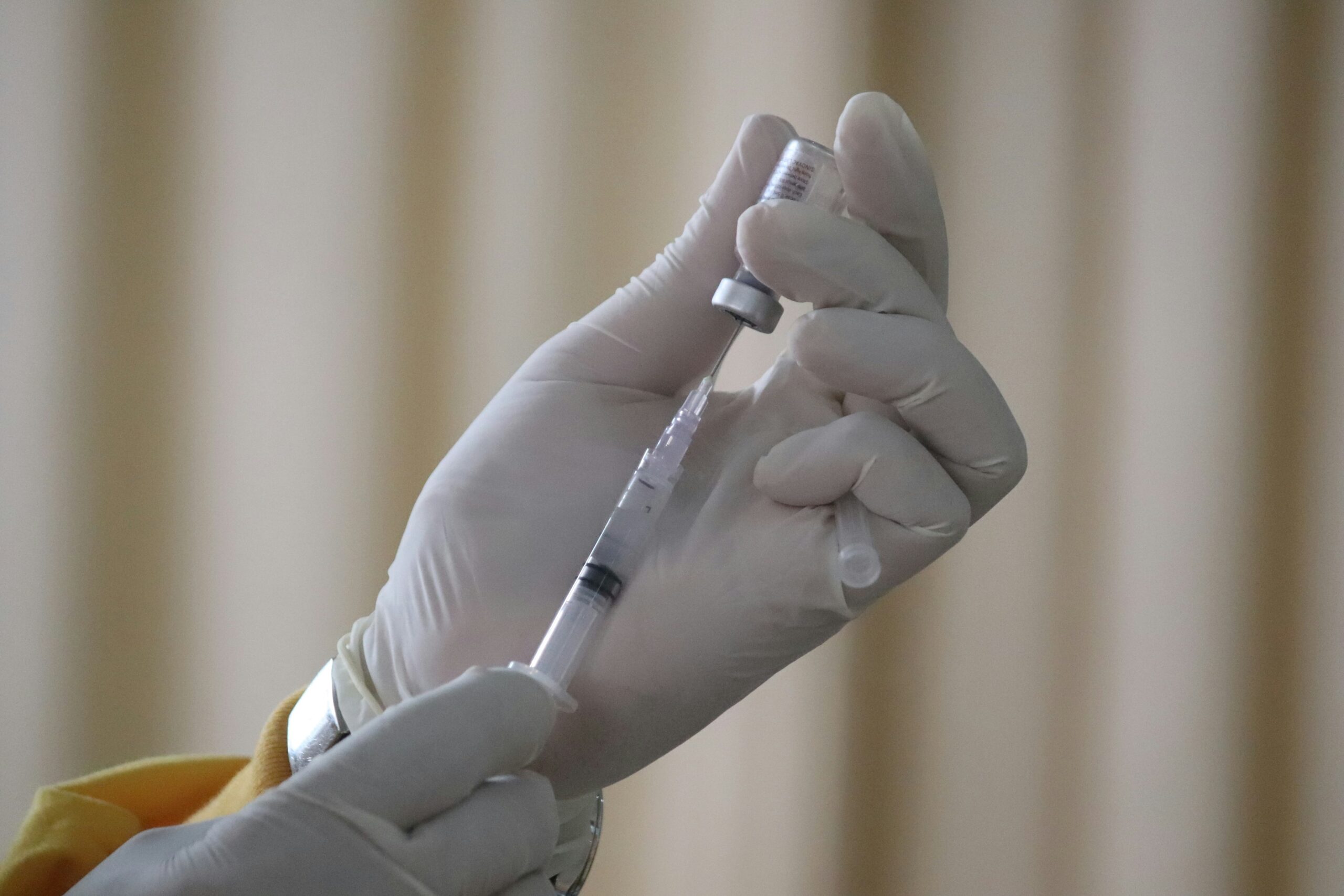A gloved hand holds a vaccine vial and syringe, drawing liquid from the vial against a blurred, neutral background—highlighting the importance of measles vaccination in protecting public health.