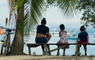 A woman and two children sit on a wooden bench by the water, facing boats on a calm bay. A bicycle leans against a palm tree nearby as sunlight filters through the leaves, capturing the peaceful spirit of spring in this relaxed scene.