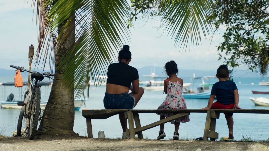 A woman and two children sit on a wooden bench by the water, facing boats on a calm bay. A bicycle leans against a palm tree nearby as sunlight filters through the leaves, capturing the peaceful spirit of spring in this relaxed scene.