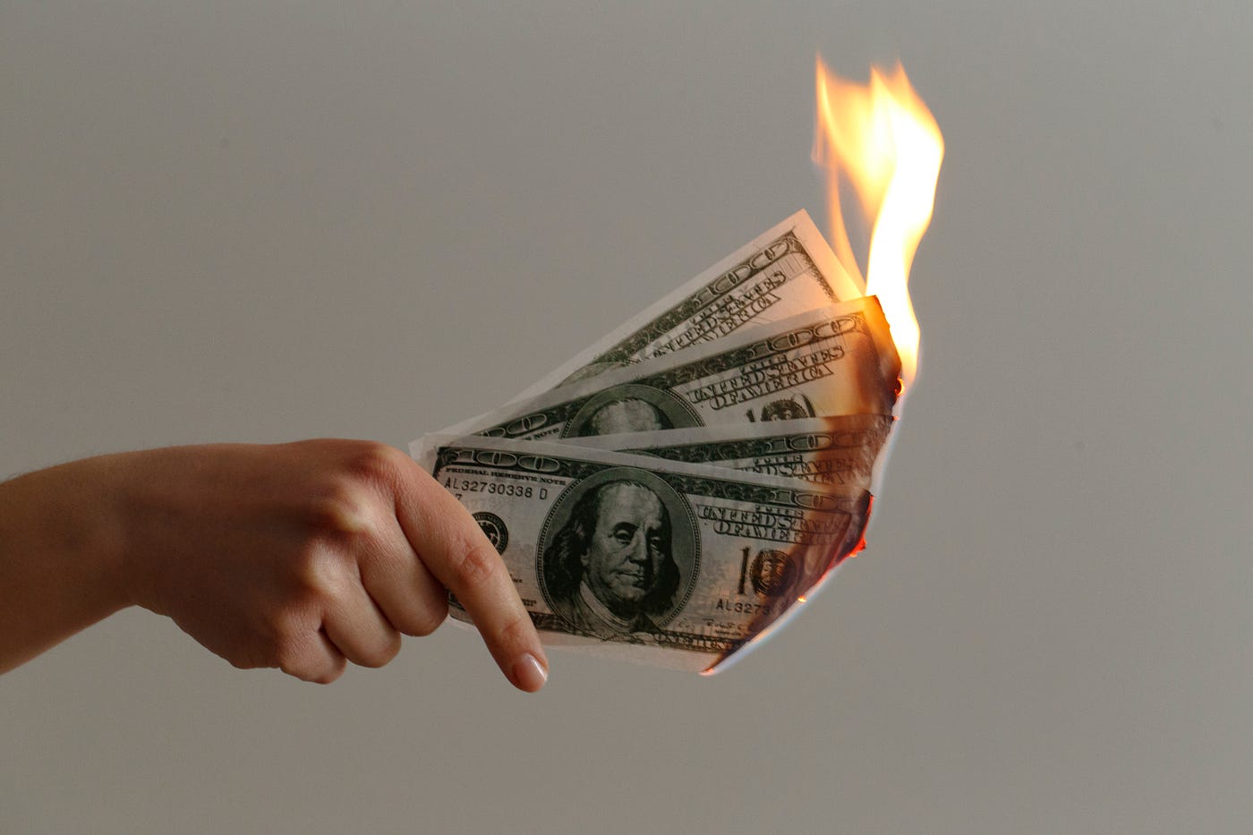 A hand holding several U.S. hundred-dollar bills, with the bills' edges on fire against a plain, light-colored background—reflecting the financial pressures of applying to a new medical school program at Waterloo.