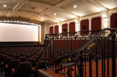 An empty, elegant movie theater with red cushioned seats and ornate walls at Princess Cinemas awaits the next July series screening. Decorative railings lead down to the seating area facing a large blank screen, ready for It Happened One Night.