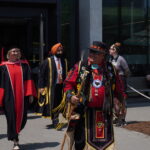 A group of people, including a person in traditional Indigenous attire and others in graduation robes, walk together outside a modern building on a sunny day, celebrating June convocation for the class of 2025.
