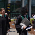 Two graduates in black gowns and green sashes walk outdoors at June convocation, holding diplomas. Other class of 2025 graduates and people stand nearby. A building with "PAC" and "SLC" signs is in the background on this sunny day.