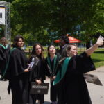 A group of graduates in black gowns and green stoles pose for a selfie outdoors during June convocation, proudly holding their diplomas, with trees, a sign, and other people in the background on a sunny day.