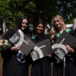 Three graduates in black gowns hold bouquets and University of Waterloo diplomas, smiling outdoors on a sunny June convocation day as the class of 2025 celebrates with trees and people in the background.