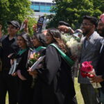 A group of graduates in black gowns with green sashes pose and smile outdoors for their June convocation, surrounded by friends holding flowers and gifts, celebrating on a sunny day.