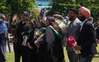 A group of graduates in black gowns with green sashes pose and smile outdoors for their June convocation, surrounded by friends holding flowers and gifts, celebrating on a sunny day.