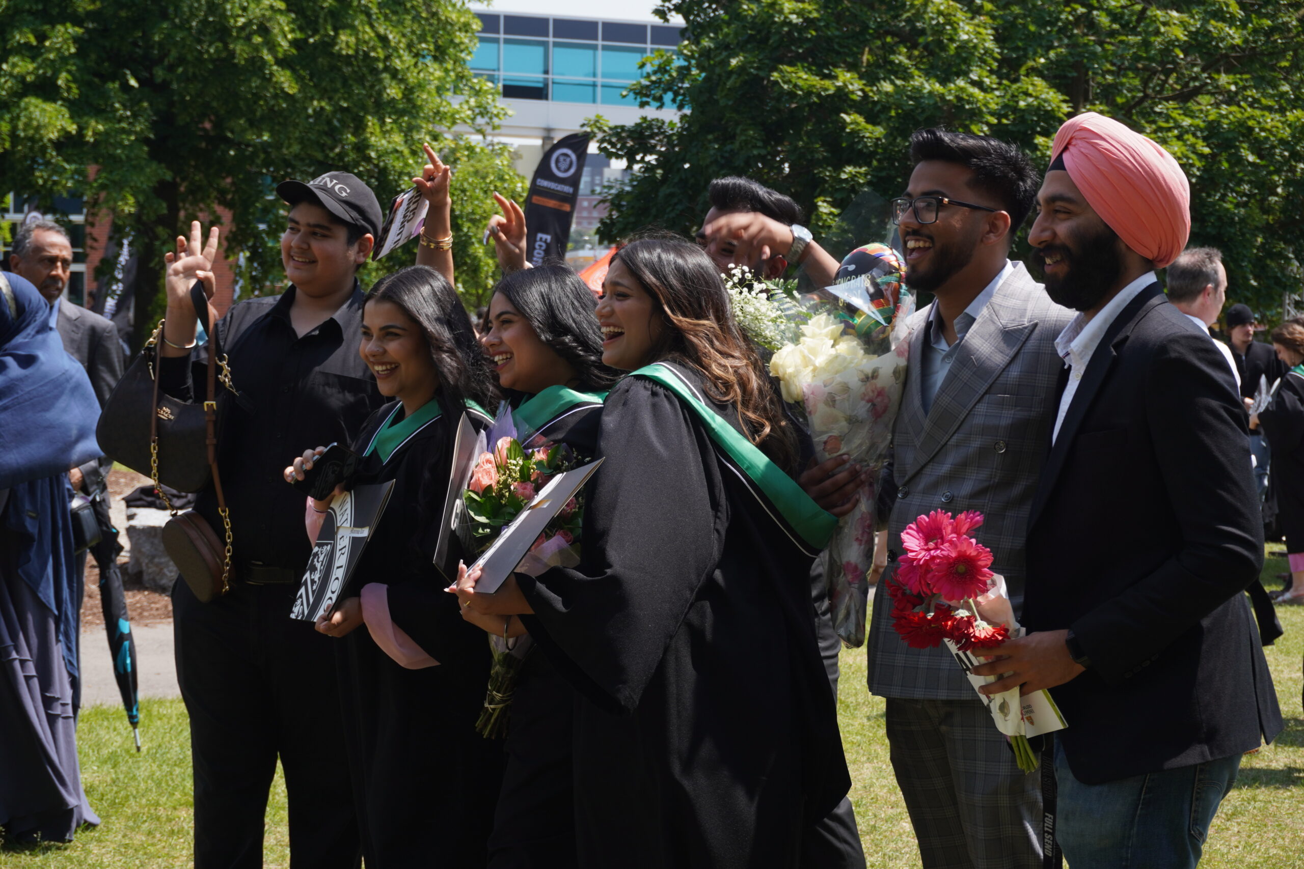 A group of graduates in black gowns with green sashes pose and smile outdoors for their June convocation, surrounded by friends holding flowers and gifts, celebrating on a sunny day.