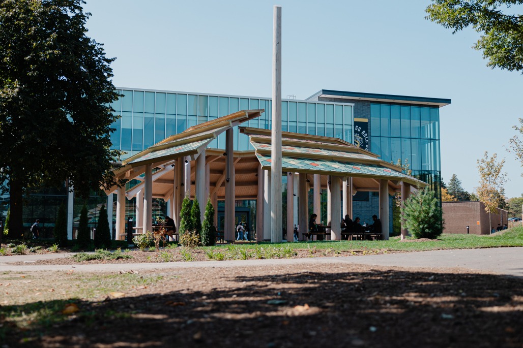 A modern glass campus building features a unique wooden pavilion in front, surrounded by trees and plants. People gather in the shaded area beneath the structure, reflecting the site’s commitment to truth and reconciliation and Indigenous initiatives.