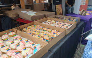 Boxes of donuts with colorful icing and small paper pride flags are arranged on a table at the Glow Centre’s Queer Life Fair at UW, with people and informational booths visible in the background.