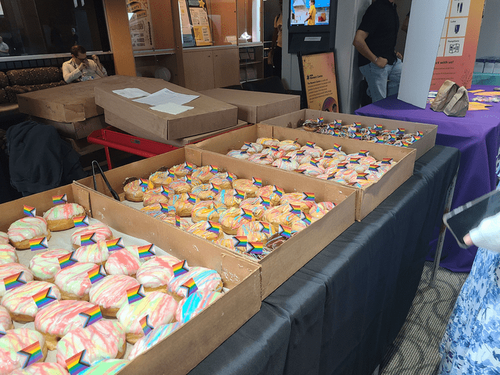 Boxes of donuts with colorful icing and small paper pride flags are arranged on a table at the Glow Centre’s Queer Life Fair at UW, with people and informational booths visible in the background.