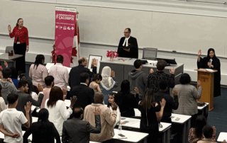 A group of people raising their right hands during a citizenship ceremony in a UW lecture hall, with officials at the front near a Canadian flag, a Faculty of Health banner, and one that reads "IMMIGRATION #CACCMPTE.