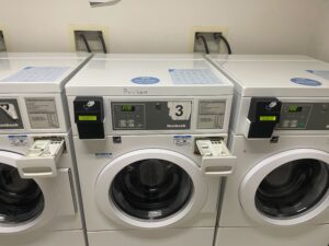 Three front-loading washing machines in a United College laundry room. The middle machine, with a handwritten “Broken” note and open detergent tray, highlights common laundry issues. All digital screens display 2:00.