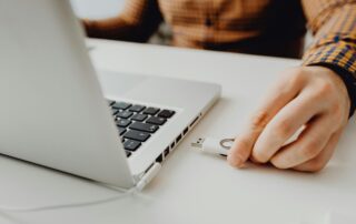 A person’s hand is about to plug a USB flash drive into a silver laptop on a white desk. The person is wearing a brown, long-sleeved, checked shirt.