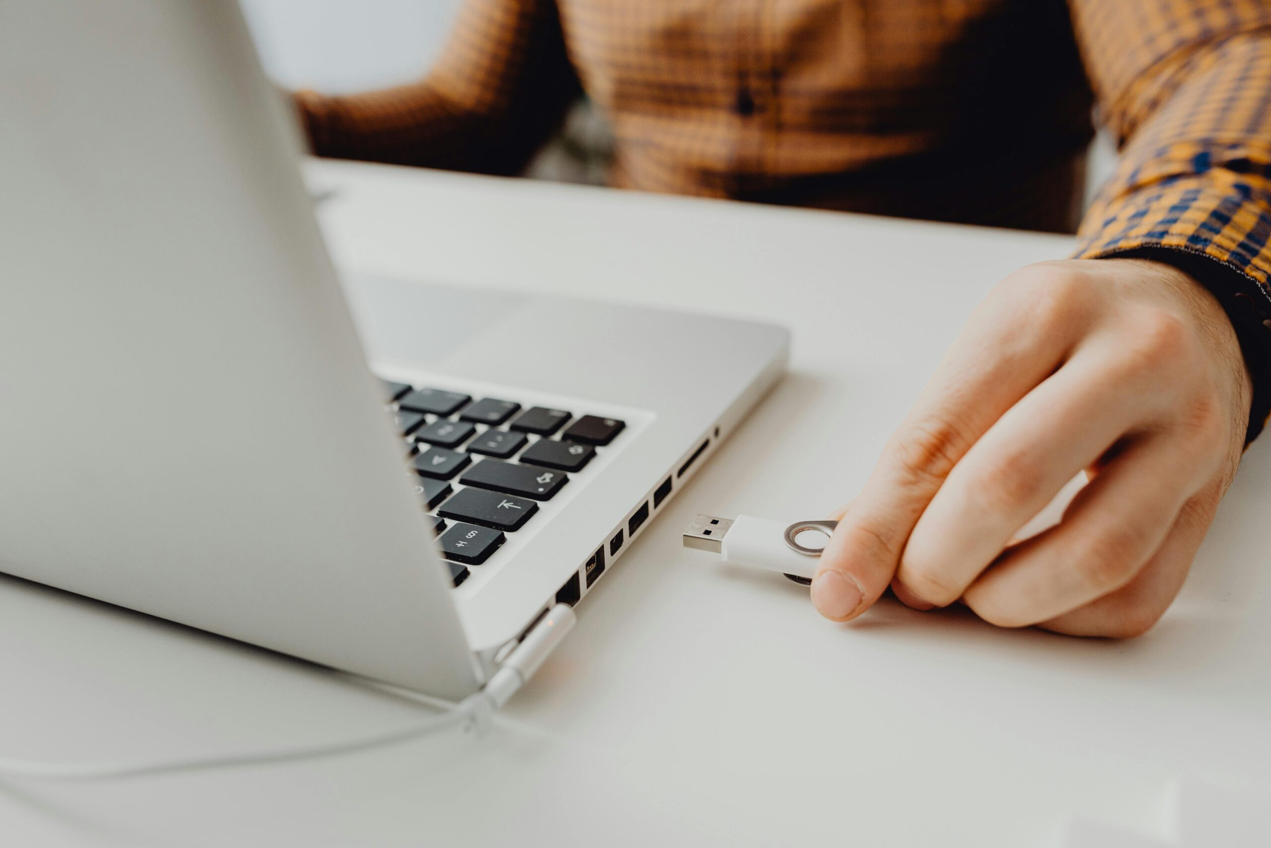 A person’s hand is about to plug a USB flash drive into a silver laptop on a white desk. The person is wearing a brown, long-sleeved, checked shirt.