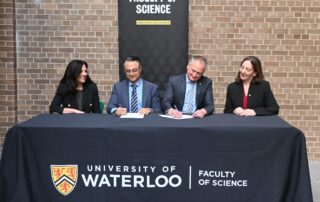 Four people sit at a table with a "University of Waterloo Faculty of Science" banner; two sign documents as others look on and smile, highlighting the university’s commitment to addressing Canada’s doctor shortage through its medical science program.