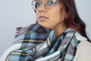 A woman with long dark hair and glasses wears a large blue and plaid scarf and a cream-colored sweater, looking slightly to the side against a plain light background.