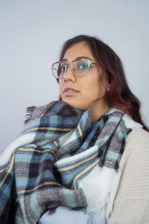 A woman with long dark hair and glasses wears a large blue and plaid scarf and a cream-colored sweater, looking slightly to the side against a plain light background.