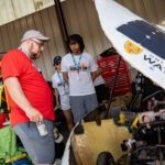 Several people stand in a workshop, examining UW Midnight Sun’s yellow solar-powered vehicle labeled "Waterloo." Tools and equipment are scattered as they inspect the car’s open cockpit—a contender for the Electrek Formula Sun Grand Prix.
