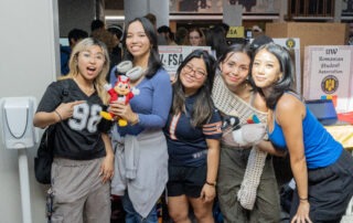 Five young women smile and pose together indoors at an event, standing in front of display tables. Showcasing free UW tools to boost your semester, they hold a red plush toy and a stuffed animal. A crowd and informational posters are visible behind them.